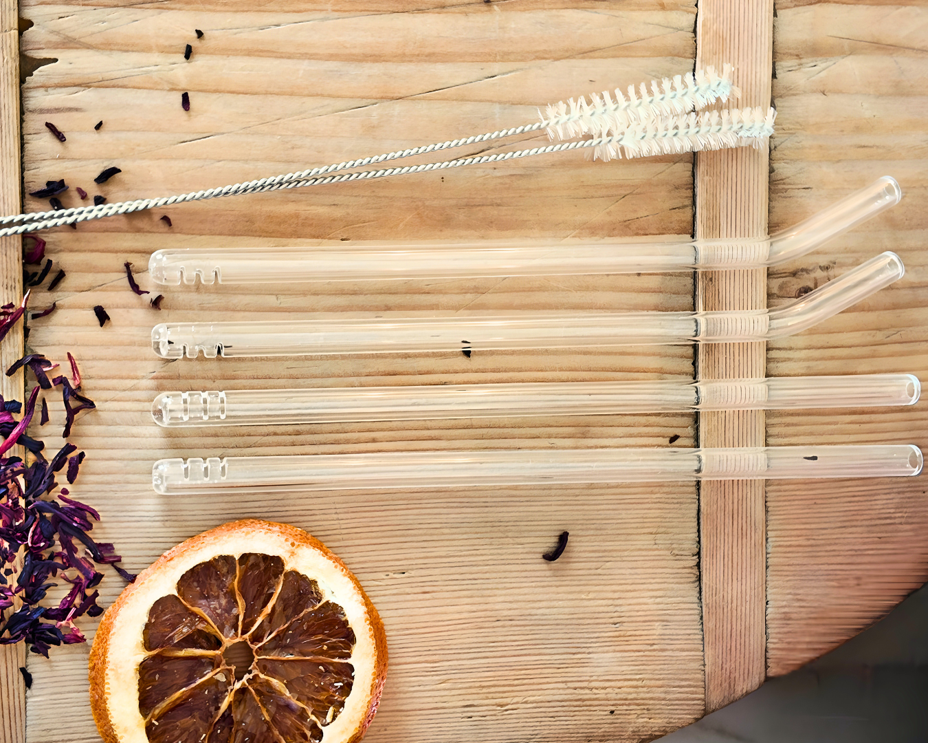 Set of filtering glass straws with a cleaning brush on a wooden surface with dried orange and flowers.